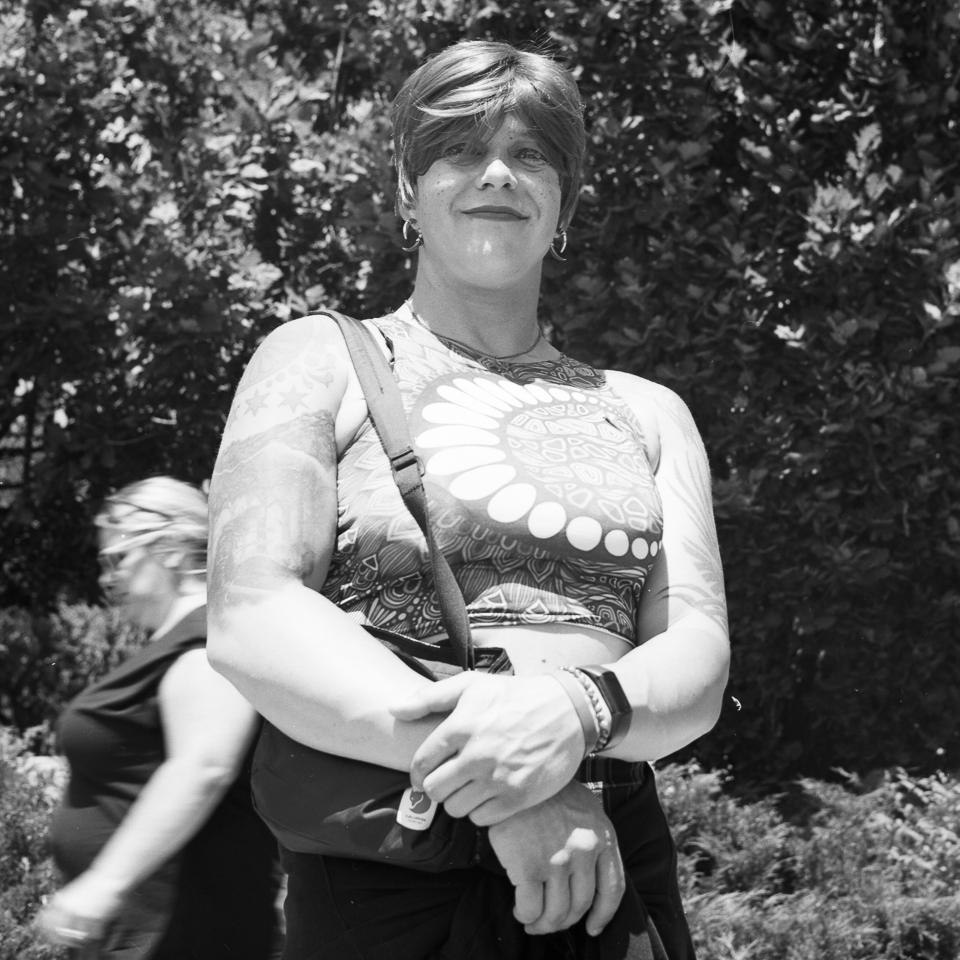 B&W portrait of a person smiling shyly at the camera, wearing a patterned shirt and holding a handbag.