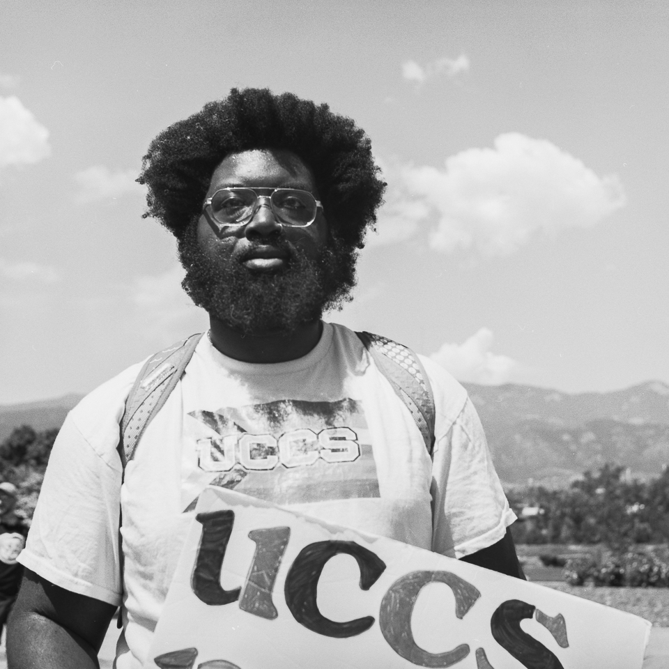 B&W portrait of a person with a beard wearing glasses wearing a UCCS t-shirt with the Pride flag as a background, and holding a UCCS poster. The foothills and clouds are in the background.